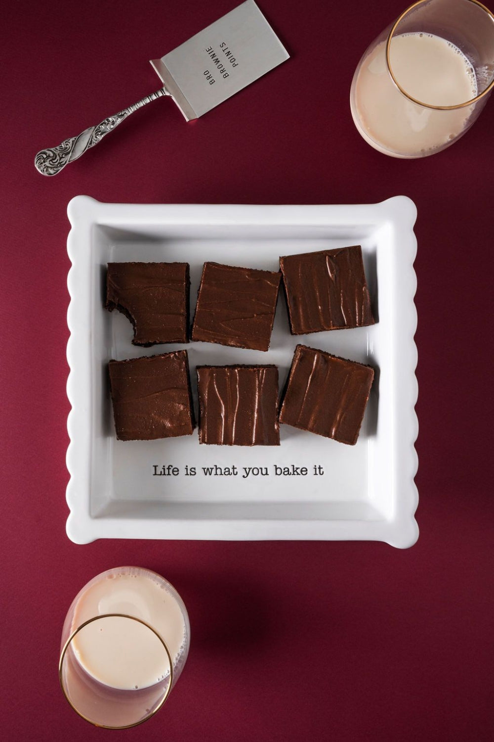 Seis brownies dentro de una caja blanca, sobre una mesa color rojo. Al lado de la caja hay dos vasos de leche y un tenedor.