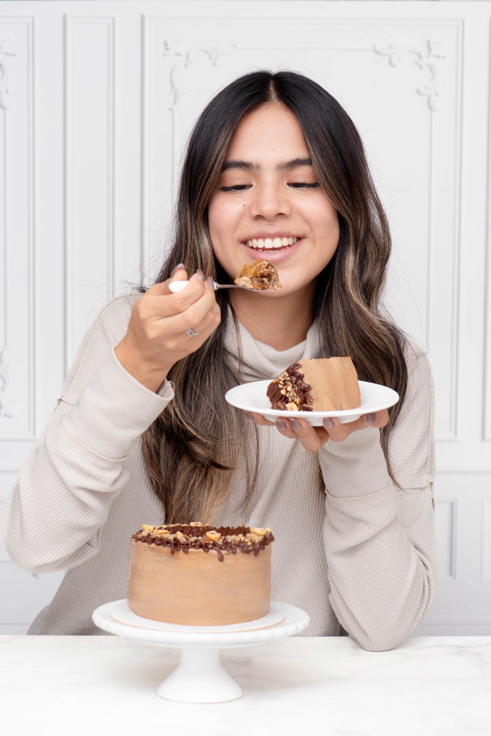 Mini torta de moka sobre un parante. Detrás de la torta hay una chica que sostiene un plato con un trozo de torta de moka en una mano y un tenedor con un pedazo del postre en la otra mano. 